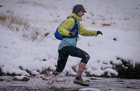 Person wearing a two tone waterproof jacket runs across a stream in a snowy lanscape