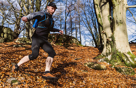Man wearing a black and grey hoodie running down hill on steep, leaf covered ground