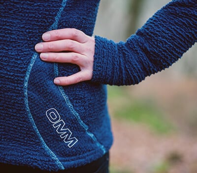 Close-up of a blue jacket with a OMM logo, worn by a person outdoors.