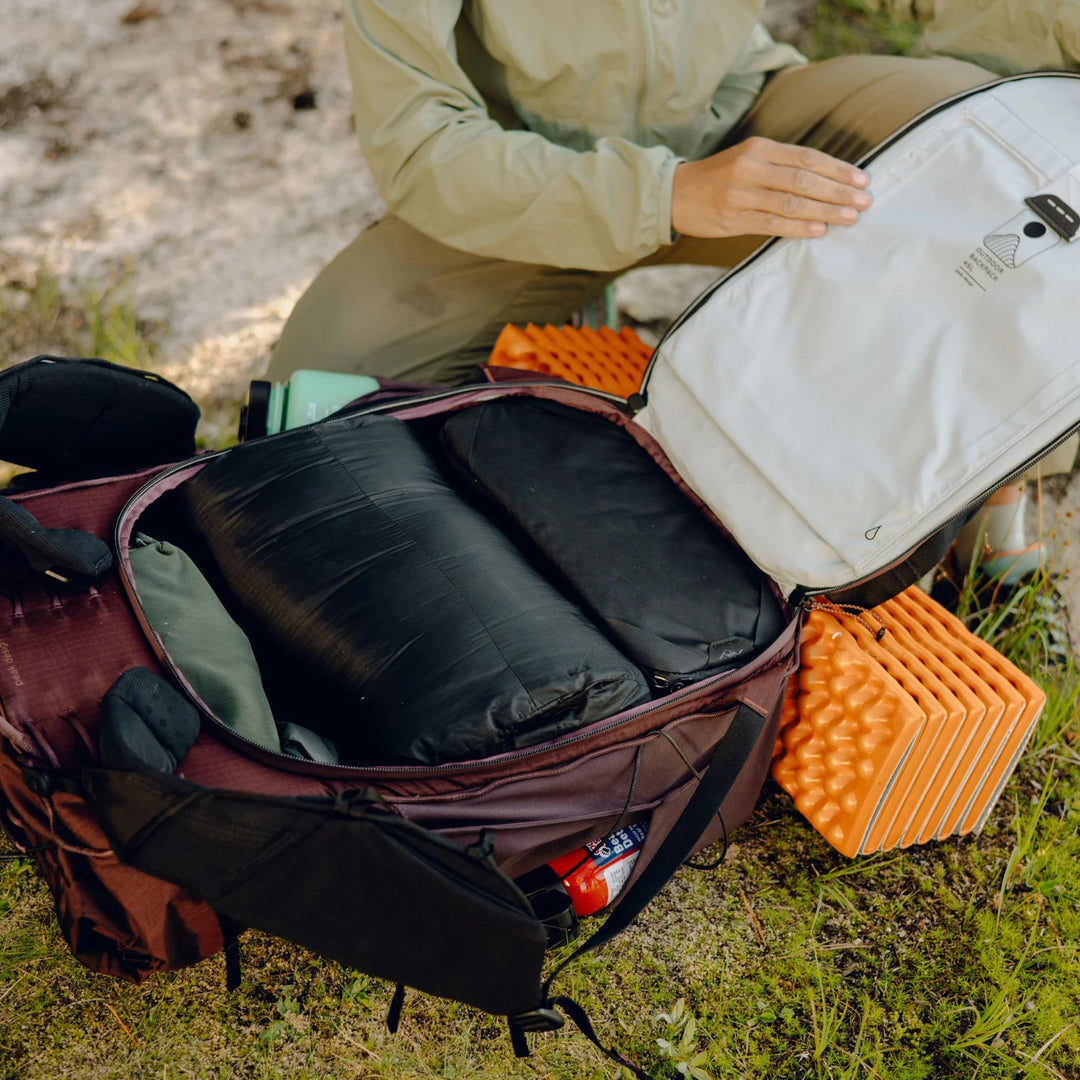 An open hiking backpack on grassy ground with a person kneeling beside it, placing or adjusting a white item inside the pack. The pack contains black sleeping bags or padded gear, and orange foam pads are visible nearby.