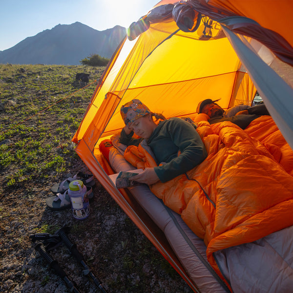 Person lying in an orange sleeping bag inside a tent with mountains in the background