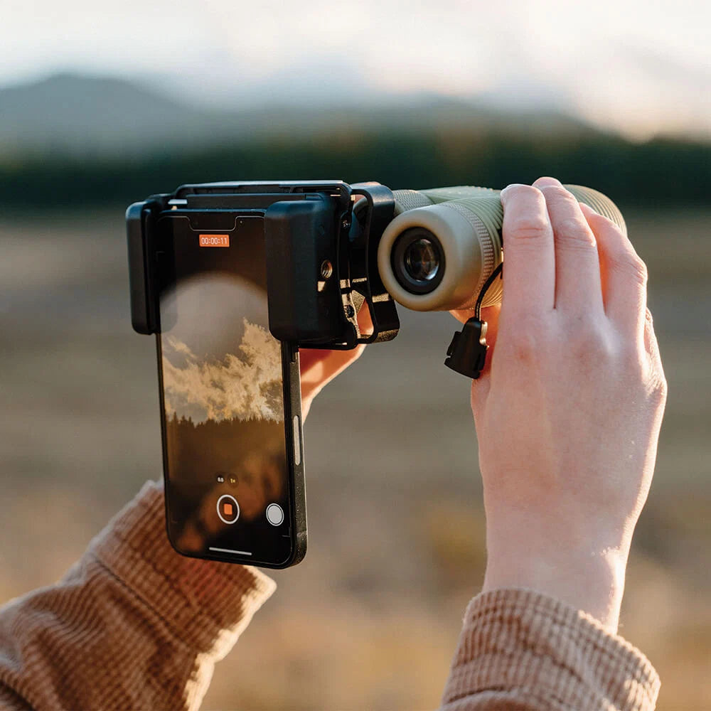 Person using binoculars with a smartphone attached to capture an image or video outdoors.