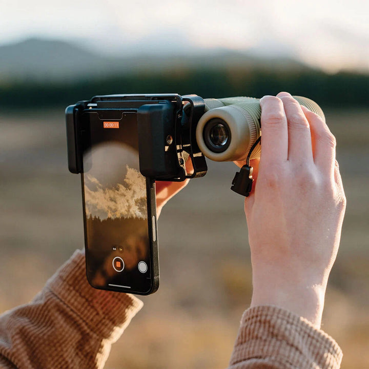 Person using binoculars with a smartphone attached to capture an image or video outdoors.