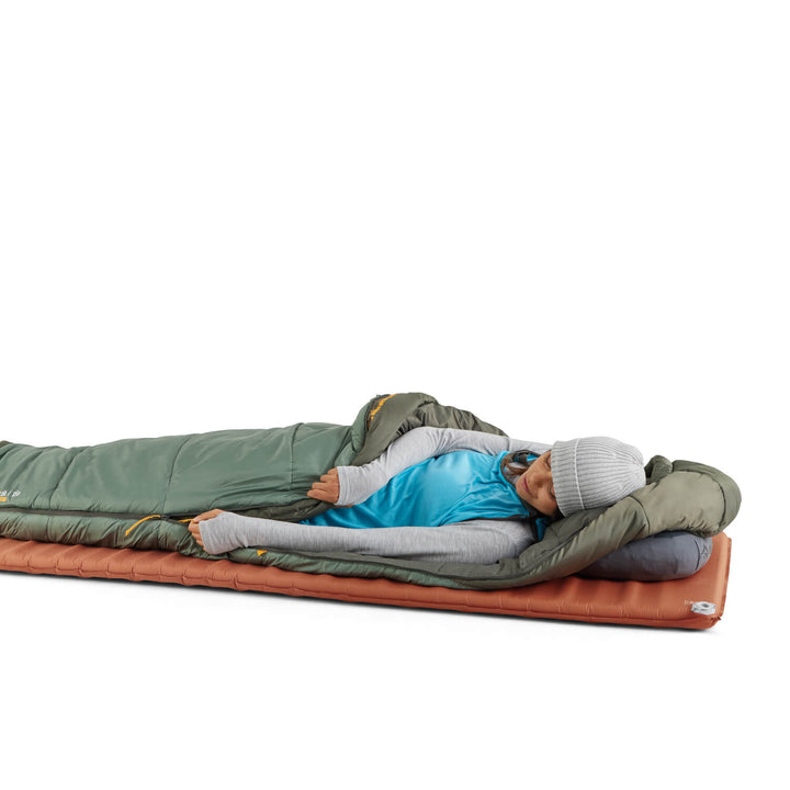 Woman zipping herself into a green sleeping bag and lying on an orange sleeping mat