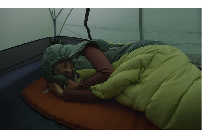 Woman in a green sleeping bag asleep on an orange sleeping mat in a twilit tense