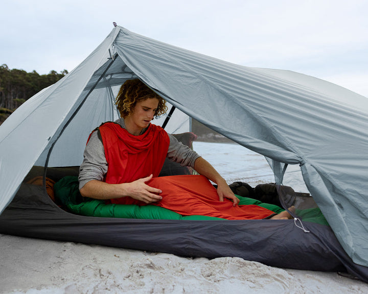 A person lying inside a tent on a beach wearing an orange sleeping bag liner