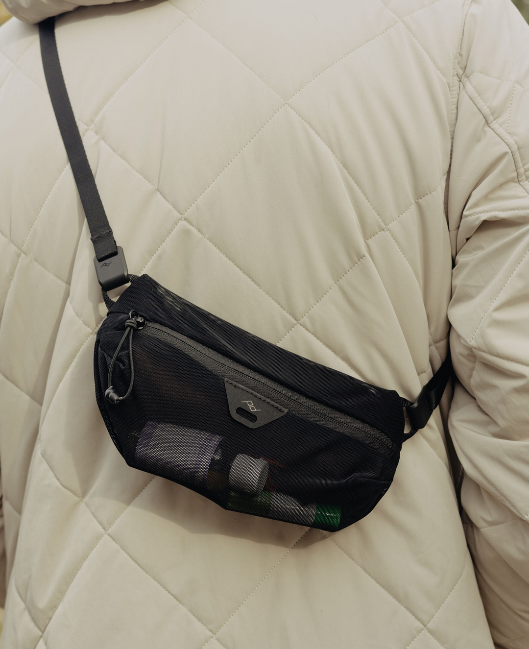 A black nylon crossbody fanny pack worn by a person in a cream quilted puffer jacket. The pouch features a front zippered pocket with a mesh window holding water bottles, a black adjustable strap, and a small silver metal logo plate.