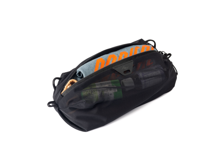A black zippered utility pouch shown open on a plain white background, with a blue and orange garment and other items partially visible inside.