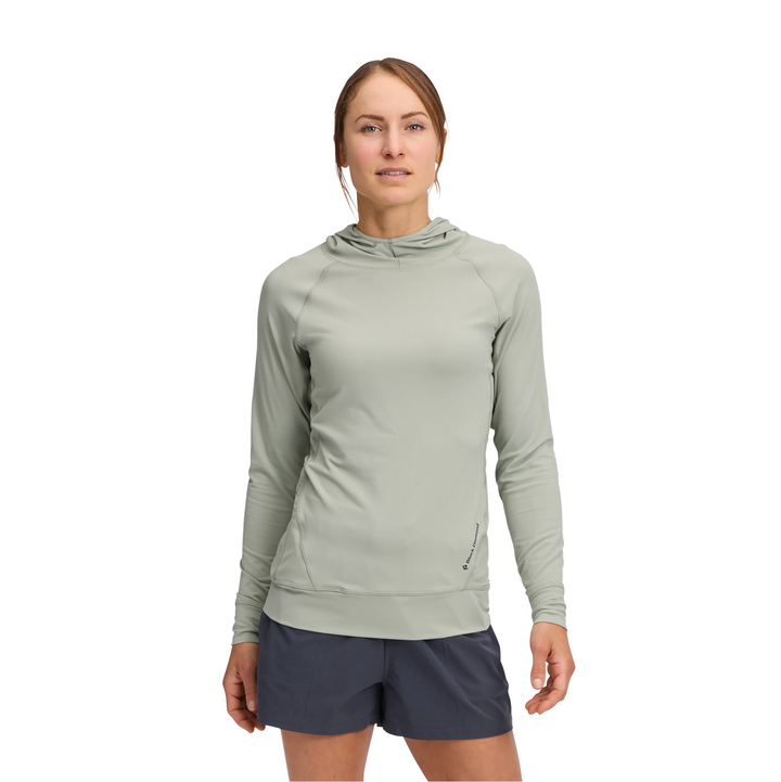 A woman modeling a light sage green hooded long-sleeve athletic top with dark navy athletic shorts, set against a plain white background.
