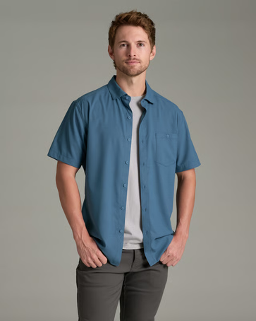 A man wearing a blue short-sleeve button-up shirt over a white t-shirt and gray pants, standing against a solid gray studio background.