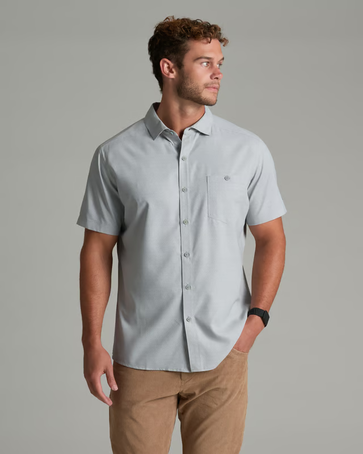 A man wearing a light gray short-sleeve button-down shirt and tan pants, standing against a solid gray studio background.