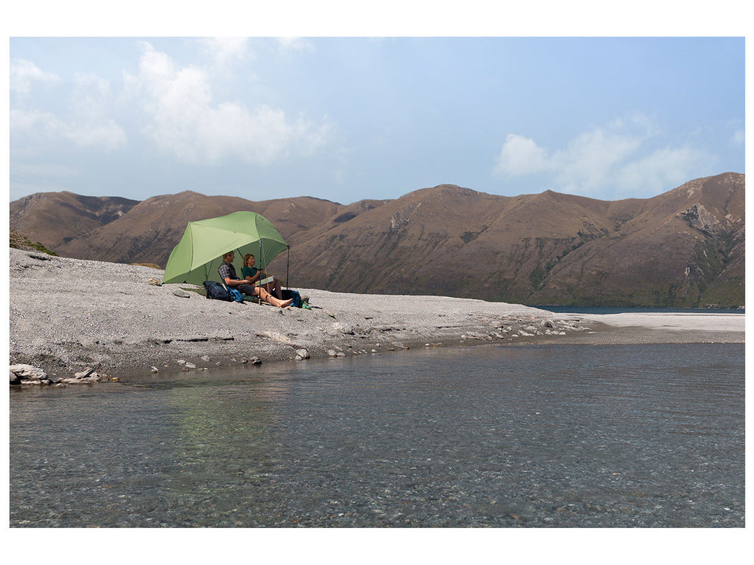Two people under a green tent fly used as a sunshade by a lake with mountains in the background