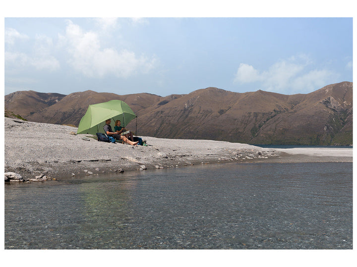 Two people under a green tent fly used as a sunshade by a lake with mountains in the background