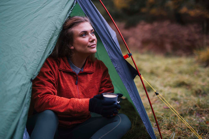 Person sitting inside a tent holding a cup while wearing gloves in a natural outdoor setting