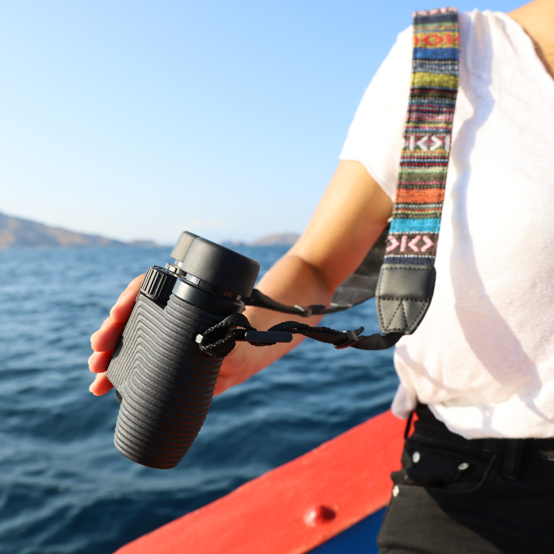 Person holding a black binocular with a colorful  woven strap, a body of water in the backhground