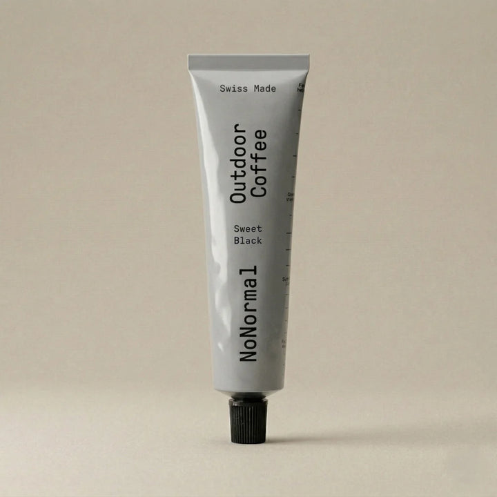A light gray squeeze tube of NoNormal Outdoor Coffee Sweet Black, standing upright on a matching light beige background.