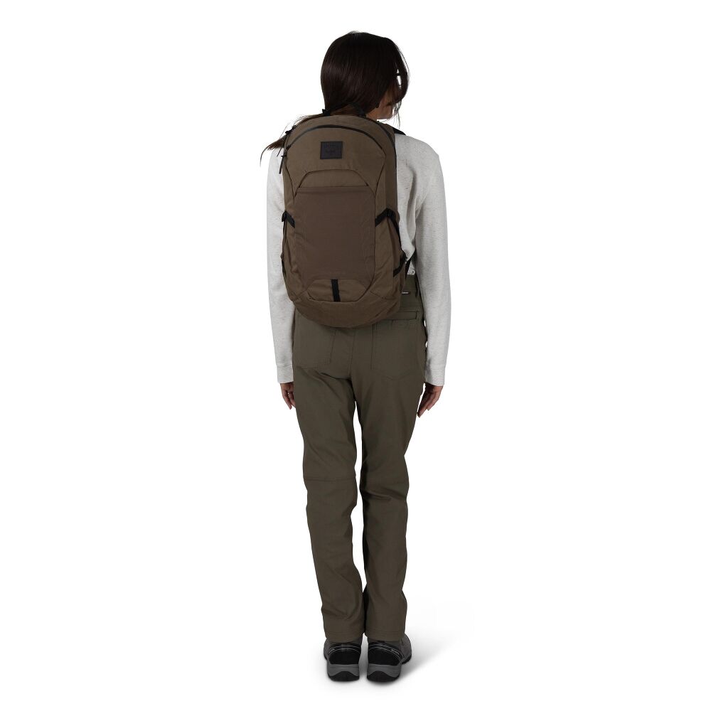 A woman viewed from behind wearing a brown backpack, olive green pants, and a light gray long-sleeve shirt against a plain white background.