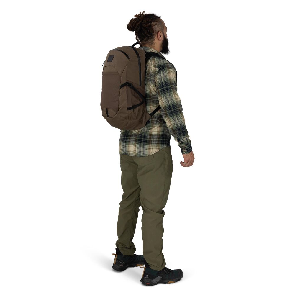A man wearing a brown backpack, a green and black plaid flannel shirt, olive green pants, and black hiking boots, shown from a rear three-quarter view against a plain white background.