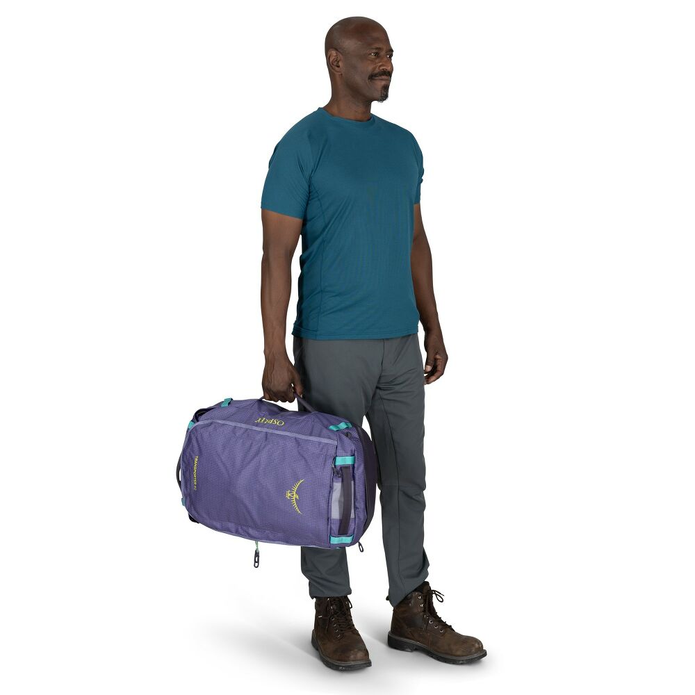 A man wearing a teal t-shirt, gray pants, and brown hiking boots carries a purple Marmot duffel bag with turquoise accents against a white background.