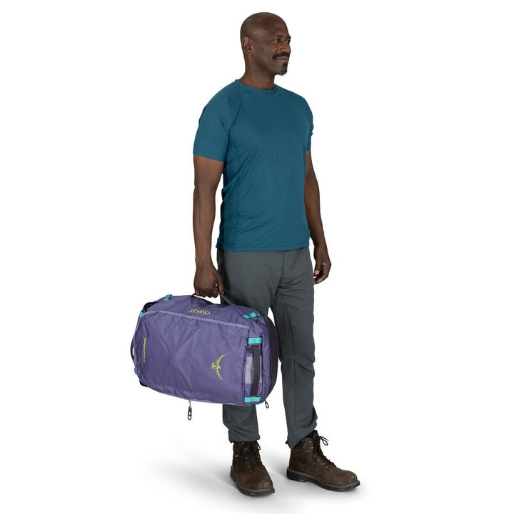 A man wearing a teal t-shirt, gray pants, and brown hiking boots carries a purple Marmot duffel bag with turquoise accents against a white background.