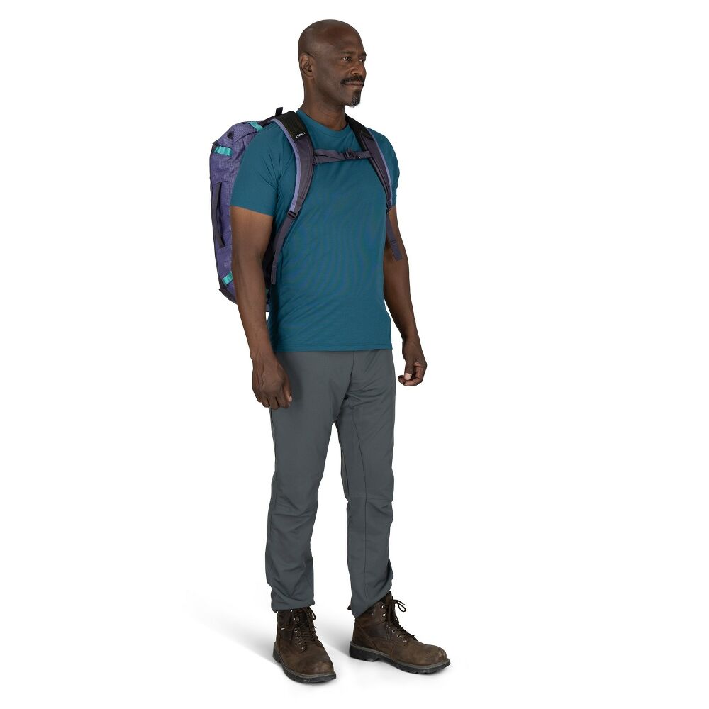 Man wearing a teal shirt, gray pants, and brown boots with a purple backpack on a white background