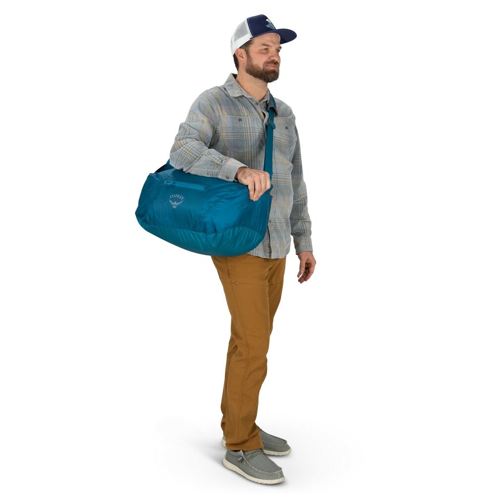 Man wearing plaid jacket and baseball cap holding a blue duffel bag over his shoulder.