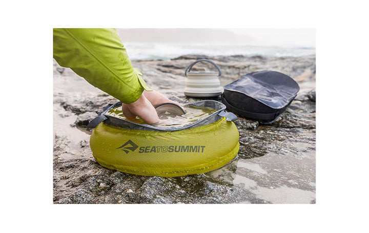 A person washing dishes in a fabric bucket on the beach