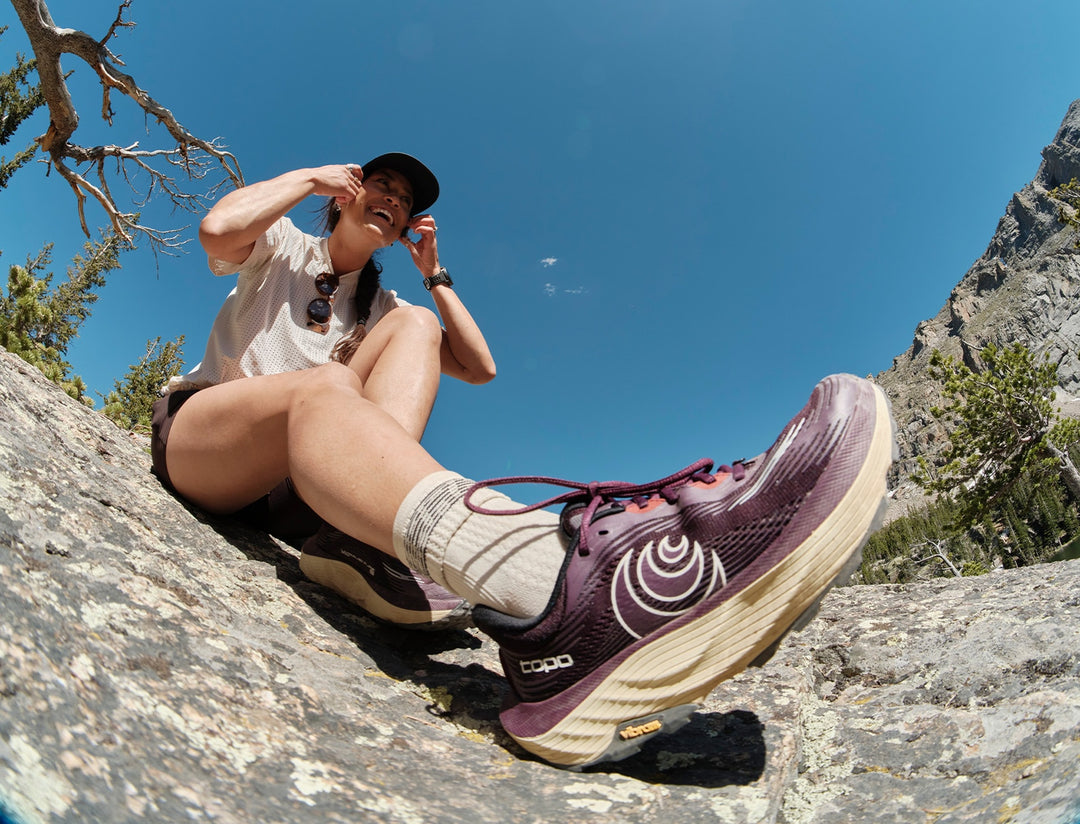 A woman sitting on a rocky mountain slope wearing a burgundy and beige trail running shoe, with the brand name &