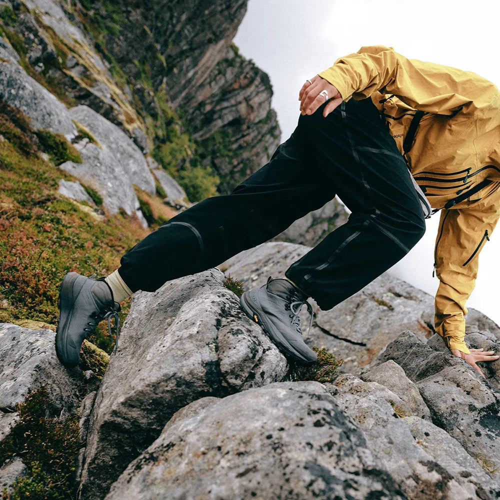 A person climbing rocky mountainside wearing black technical pants, gray hiking boots, and a yellow jacket, with steep rock formations and vegetation in the background.