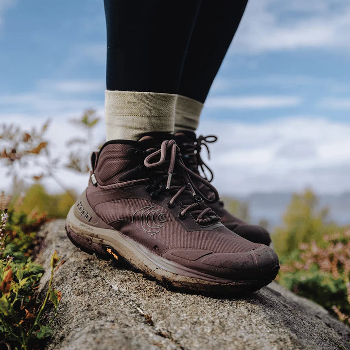 A close-up of a person wearing brown mid-cut hiking boots with black leggings, standing on a large rock outdoors with a blurred natural background.