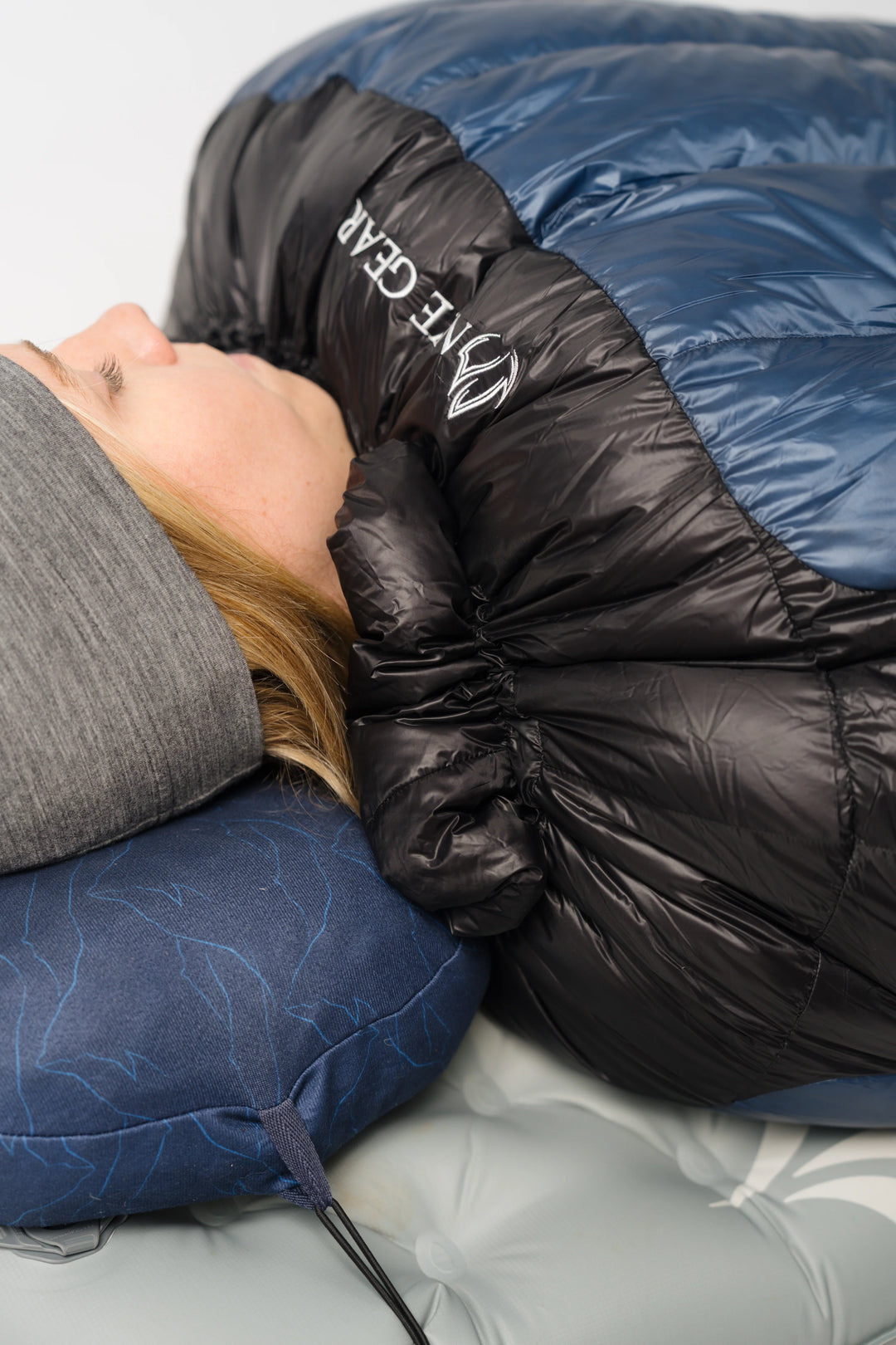 A close-up lifestyle shot of a person sleeping in a puffer sleeping bag. The sleeping bag is dark blue and black with the brand name &