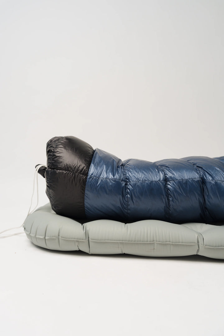 A blue and black puffer sleeping bag laid flat on a light gray sleeping pad against a plain white studio background.