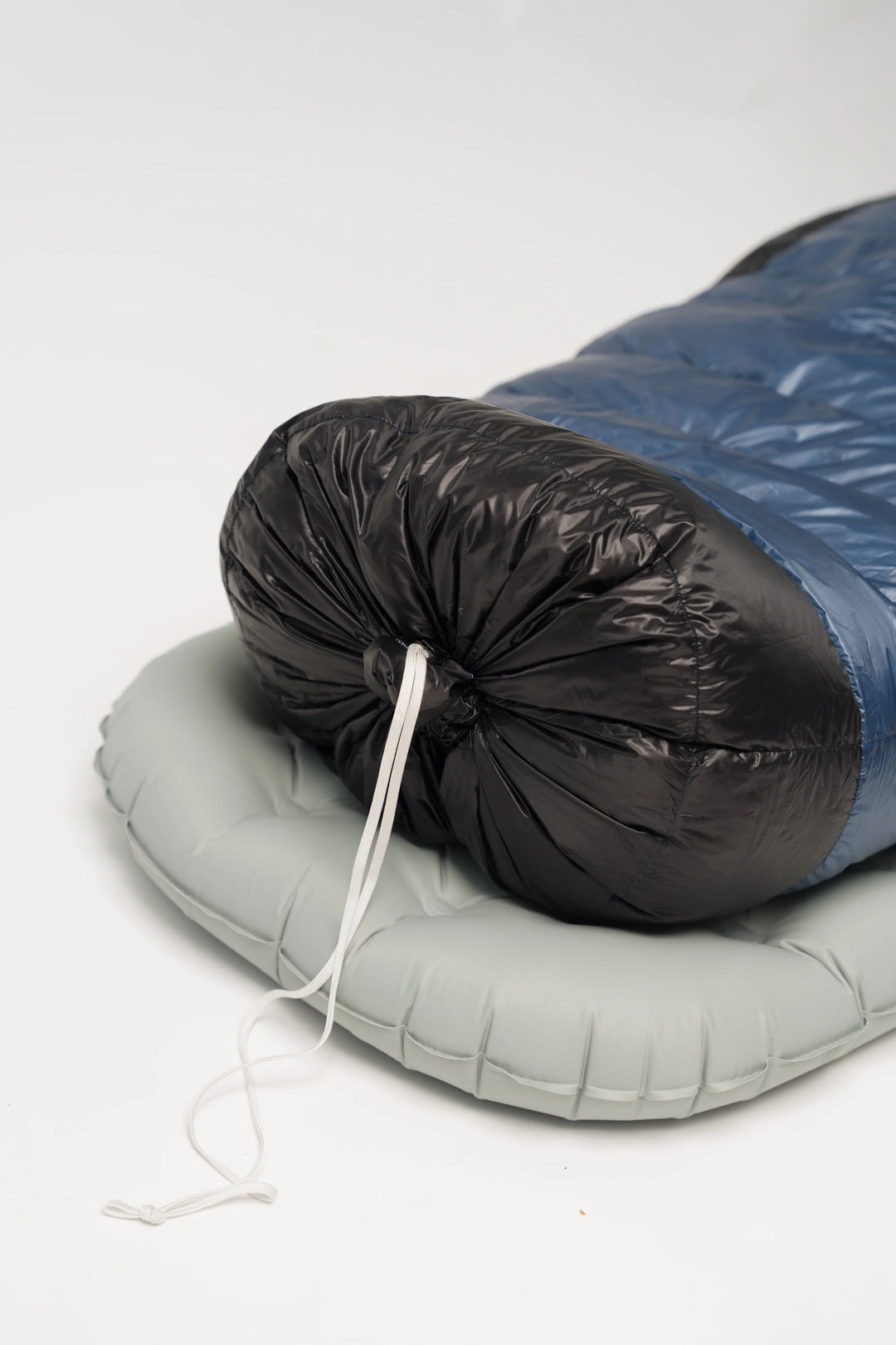A packed sleeping bag with a black insulated sleeping pouch and blue outer shell, resting on a light gray inflatable pad against a white background.