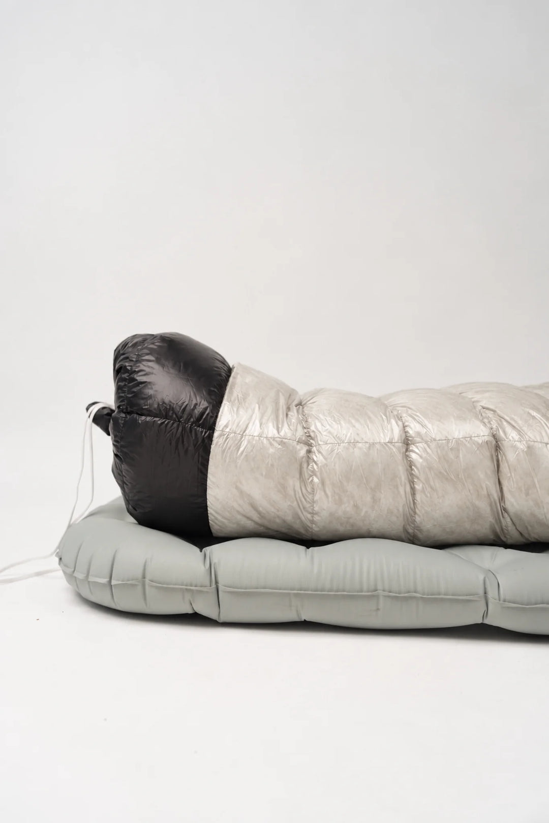 A silver puffer sleeping bag with a black hood, laid flat on a light gray sleeping pad against a plain white studio background.