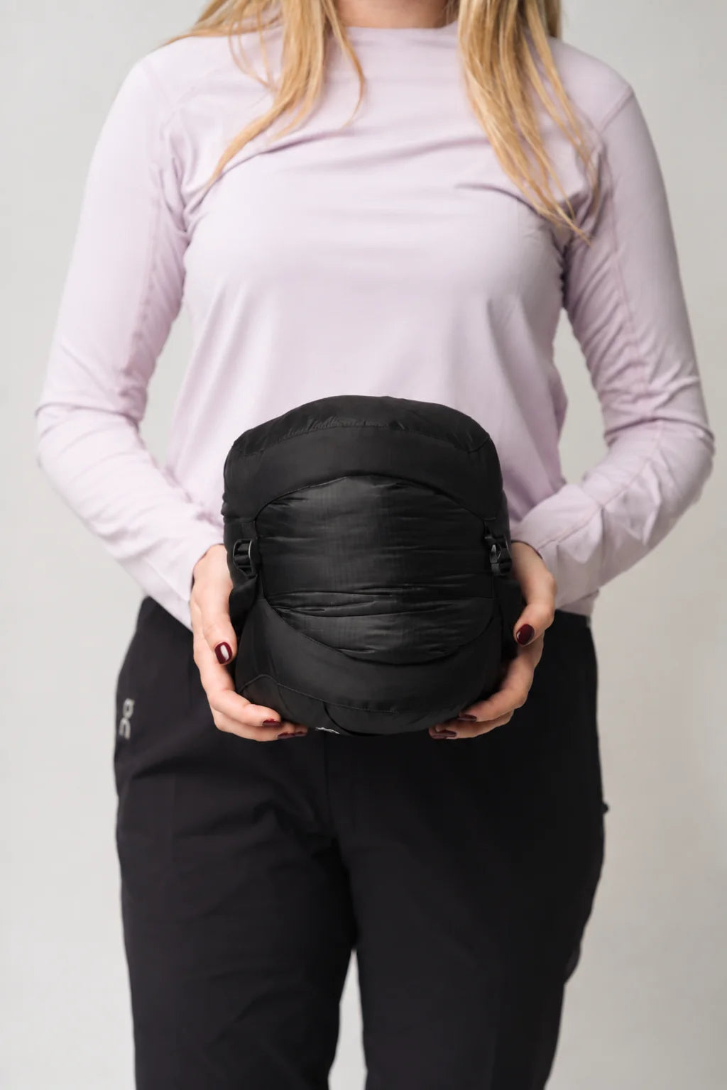 A woman in a light purple long-sleeve top and black pants holds a black, compact, rounded travel pillow or soft bag against a light gray background.