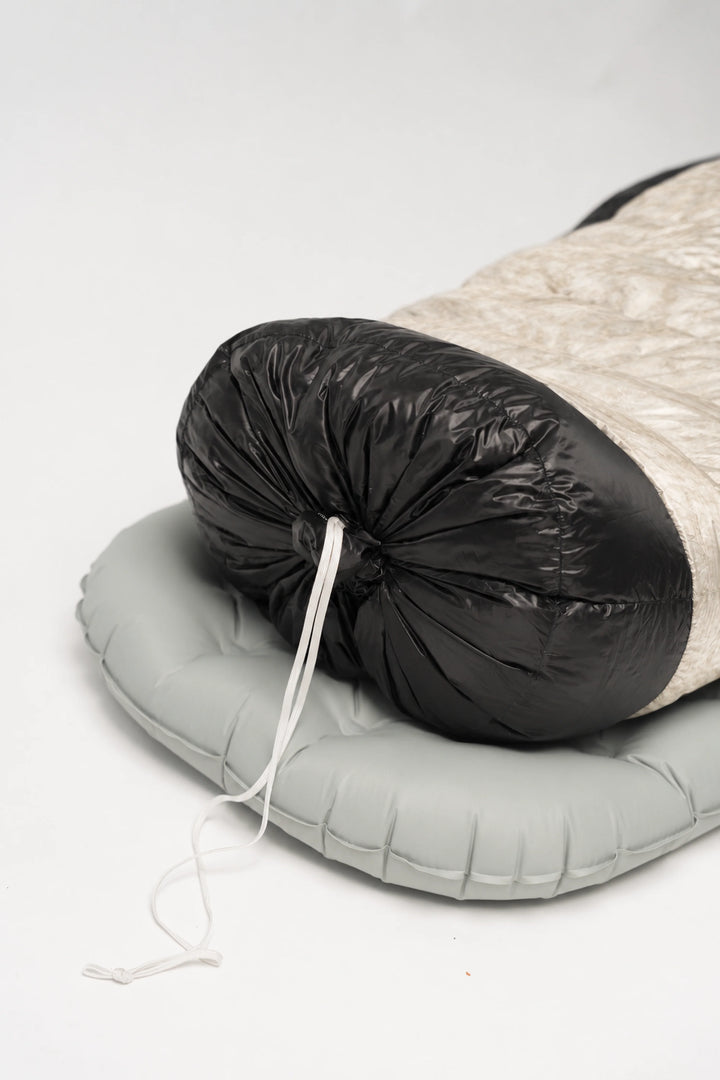 A packed sleeping bag with a black hood and a beige quilted body, resting on a light gray inflatable sleeping pad against a white studio background.