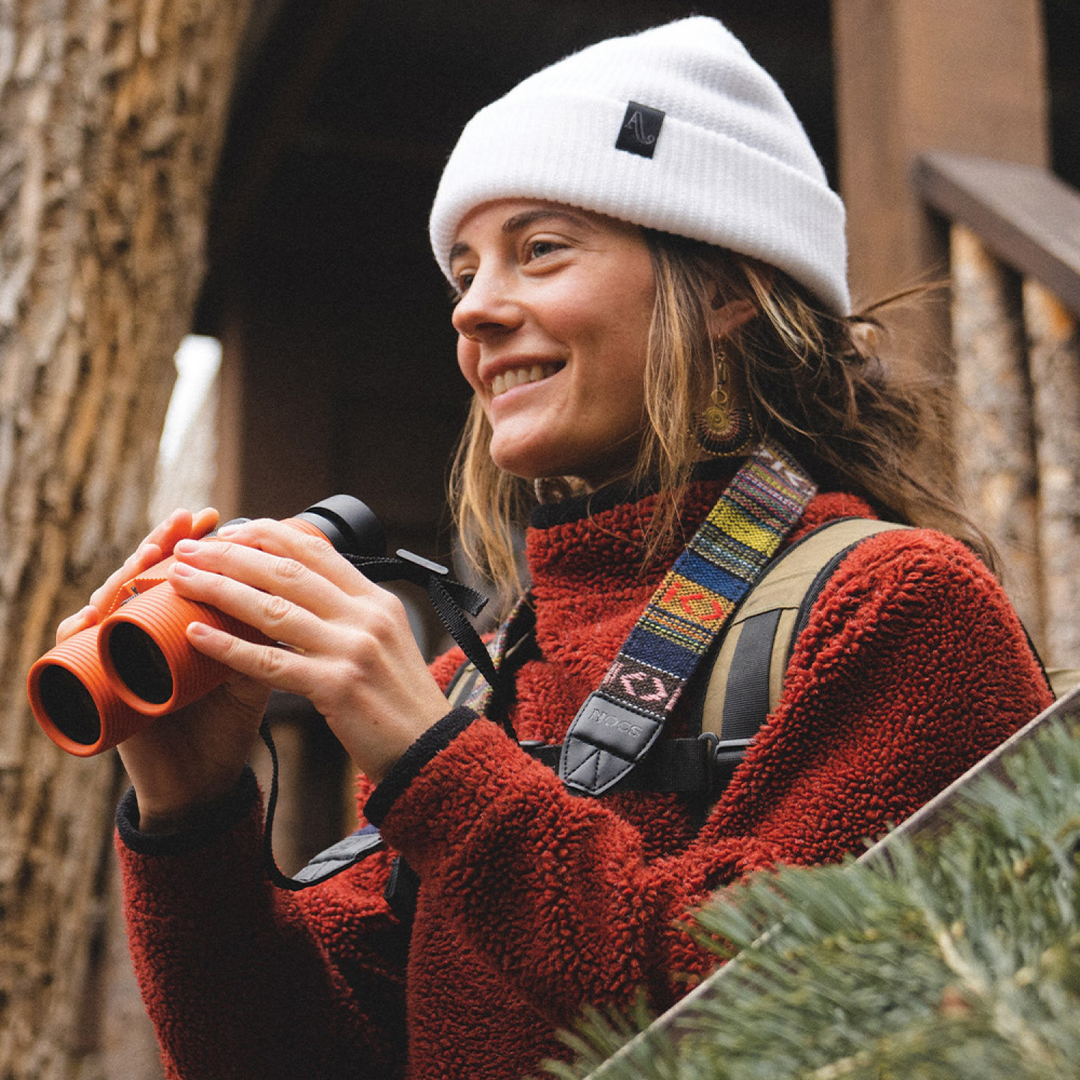 Person in a red jacket and white beanie holding orange binoculars and wearing a multicoloured camera strap.