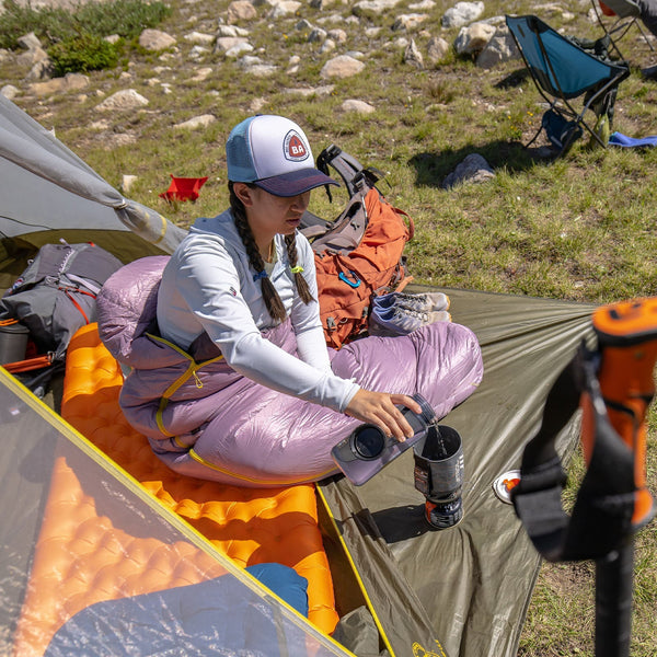 Person sitting inside a tent with camping gear around, in an outdoor setting.