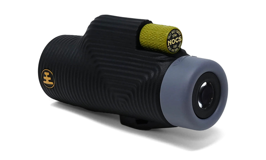 A black ribbed monocular with a gray end cap and a small yellow accent ring, photographed on a clean white background. The yellow ring shows the brand name &