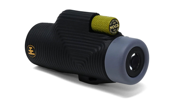 A black ribbed monocular with a gray end cap and a small yellow accent ring, photographed on a clean white background. The yellow ring shows the brand name &