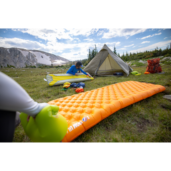 People setting up camp in an alpine meadow, inflatable orange sleeping mat center foreground of shot