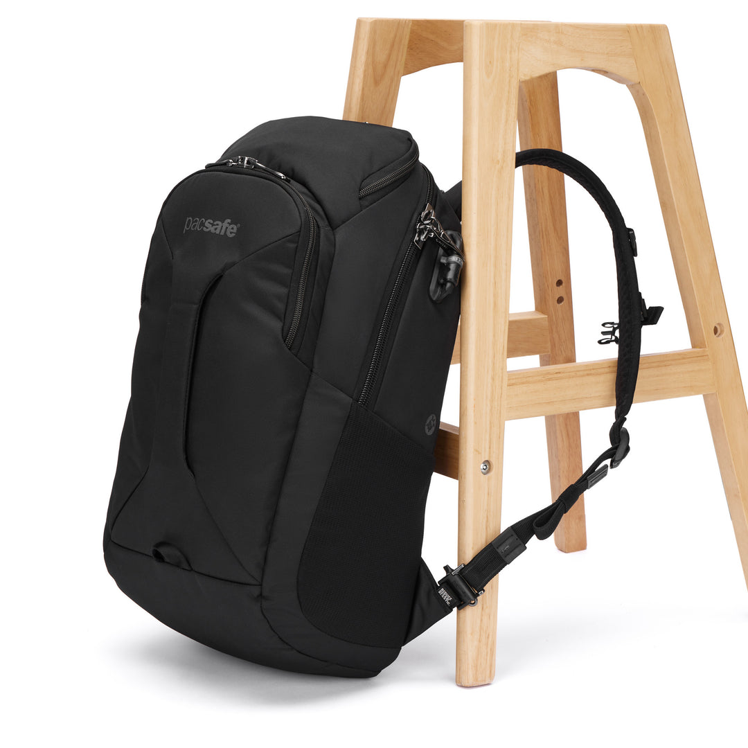 Black backpack with Pacsafe logo leaning against a wooden stool with one strap clipped through a leg