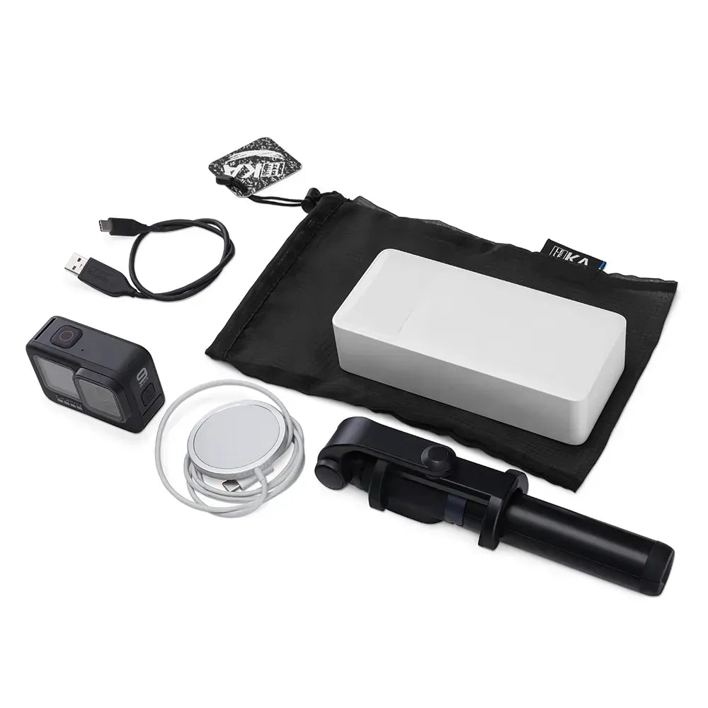 A selfie stick kit laid out on a white background, including a black selfie stick, a white rectangular accessory, a black carrying pouch, a USB cable, a small black action camera, a round silver accessory, and a small branded tag.