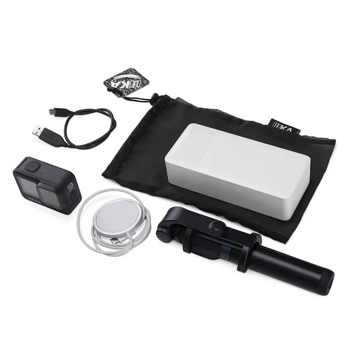 A selfie stick kit laid out on a white background, including a black selfie stick, a white rectangular accessory, a black carrying pouch, a USB cable, a small black action camera, a round silver accessory, and a small branded tag.