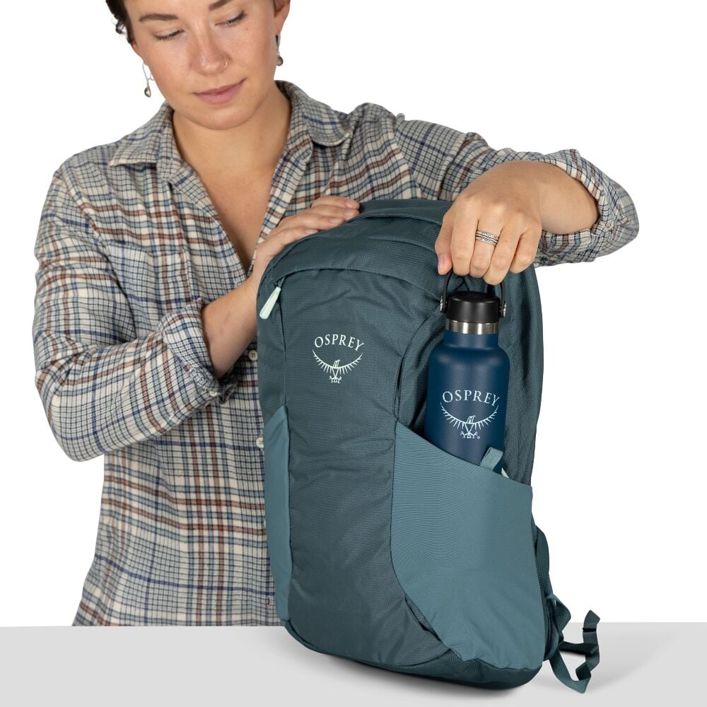 Person holding a teal Osprey backpack with a blue water bottle on a white background