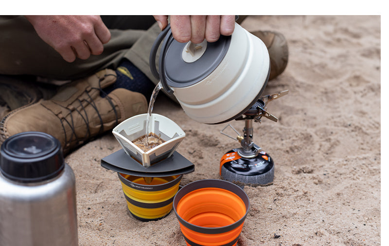 Person making coffee with a collapsible pour over, witting cross legged on sand
