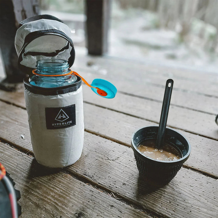 Camping setup with a water bottle in an insulated pouch, cup, and spoon on a wooden surface.