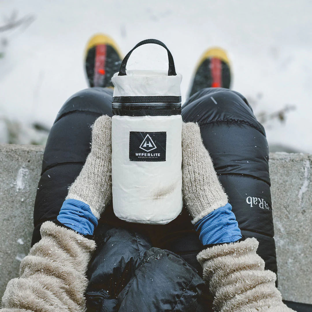 Person holding a white insulated bottle with a logo, wearing winter gloves and black pants.