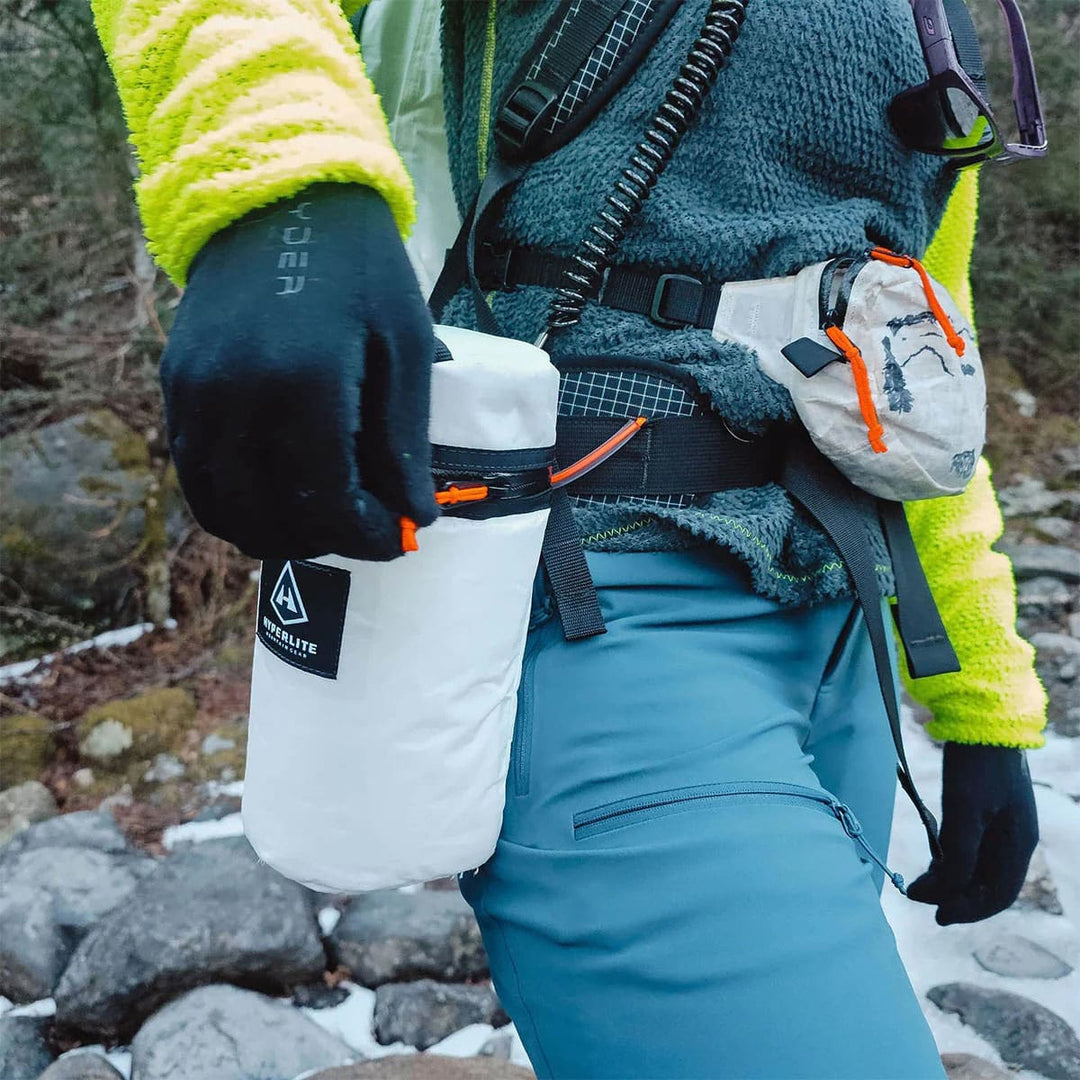 Person in outdoor gear holding a white insulated bottle with a green and yellow jacket.