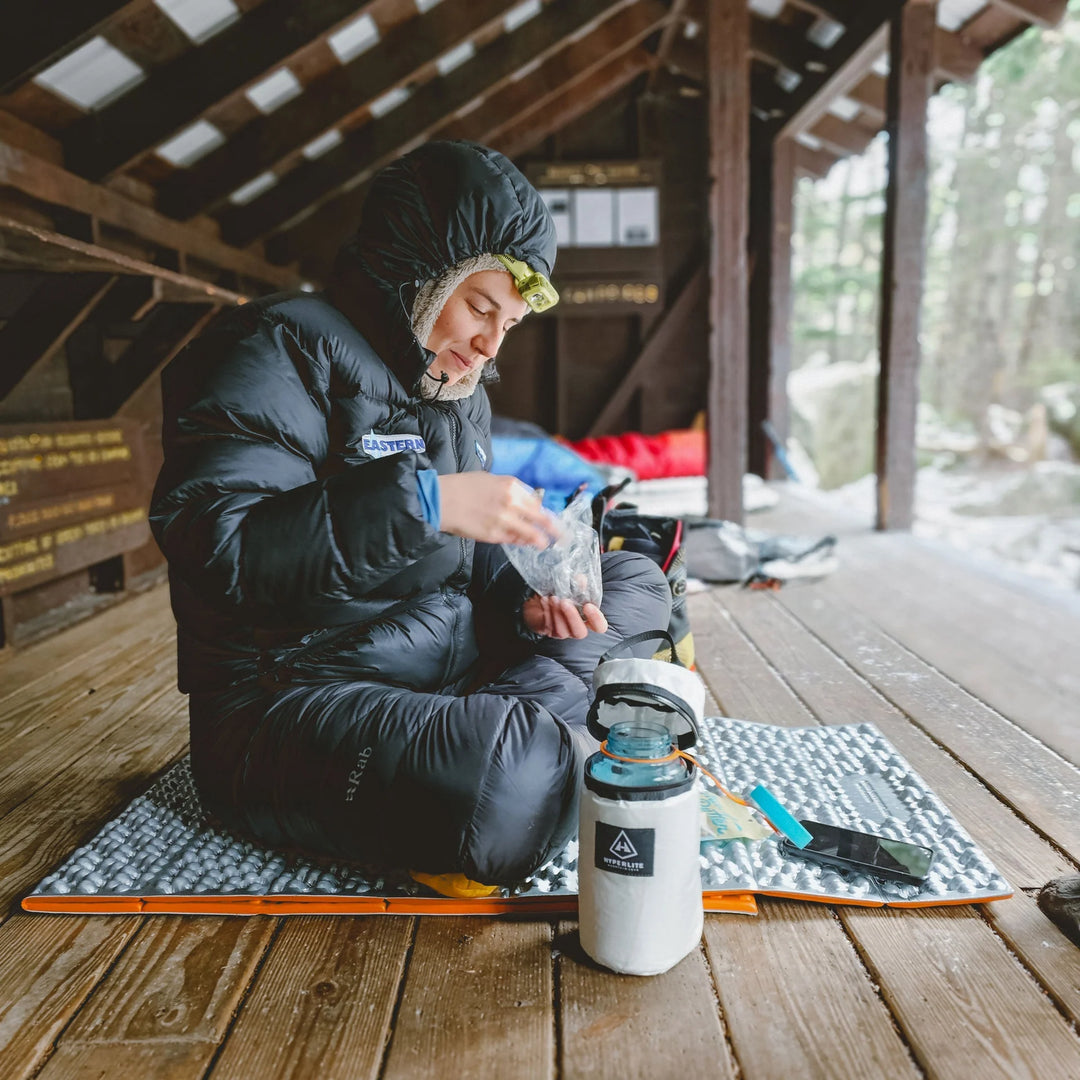 Person in winter clothing sitting on a wooden floor with camping gear, including a insulated bottle.